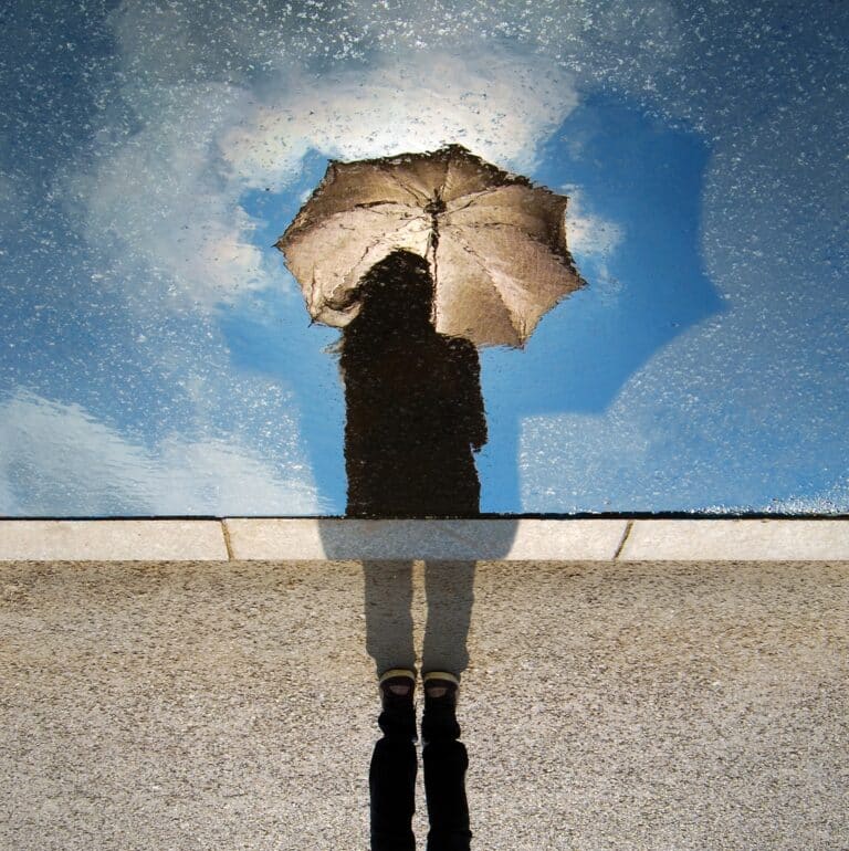 Reflection of a person holding an umbrella in a puddle, appearing as if they are standing in the sky.