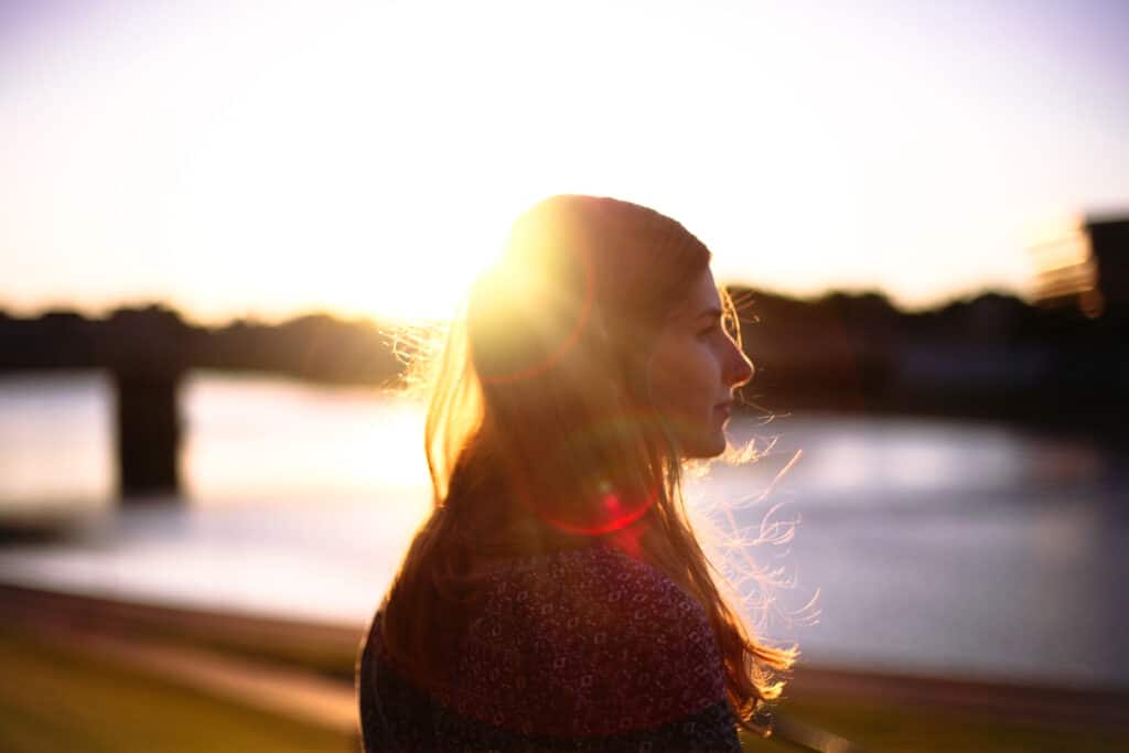 A person with long hair stands by a river at sunset, with the sun creating a bright halo effect behind them.