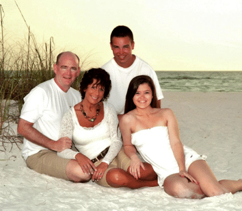 Four people posing together on a sandy beach with dunes in the background, all wearing white outfits, smiling towards the camera.