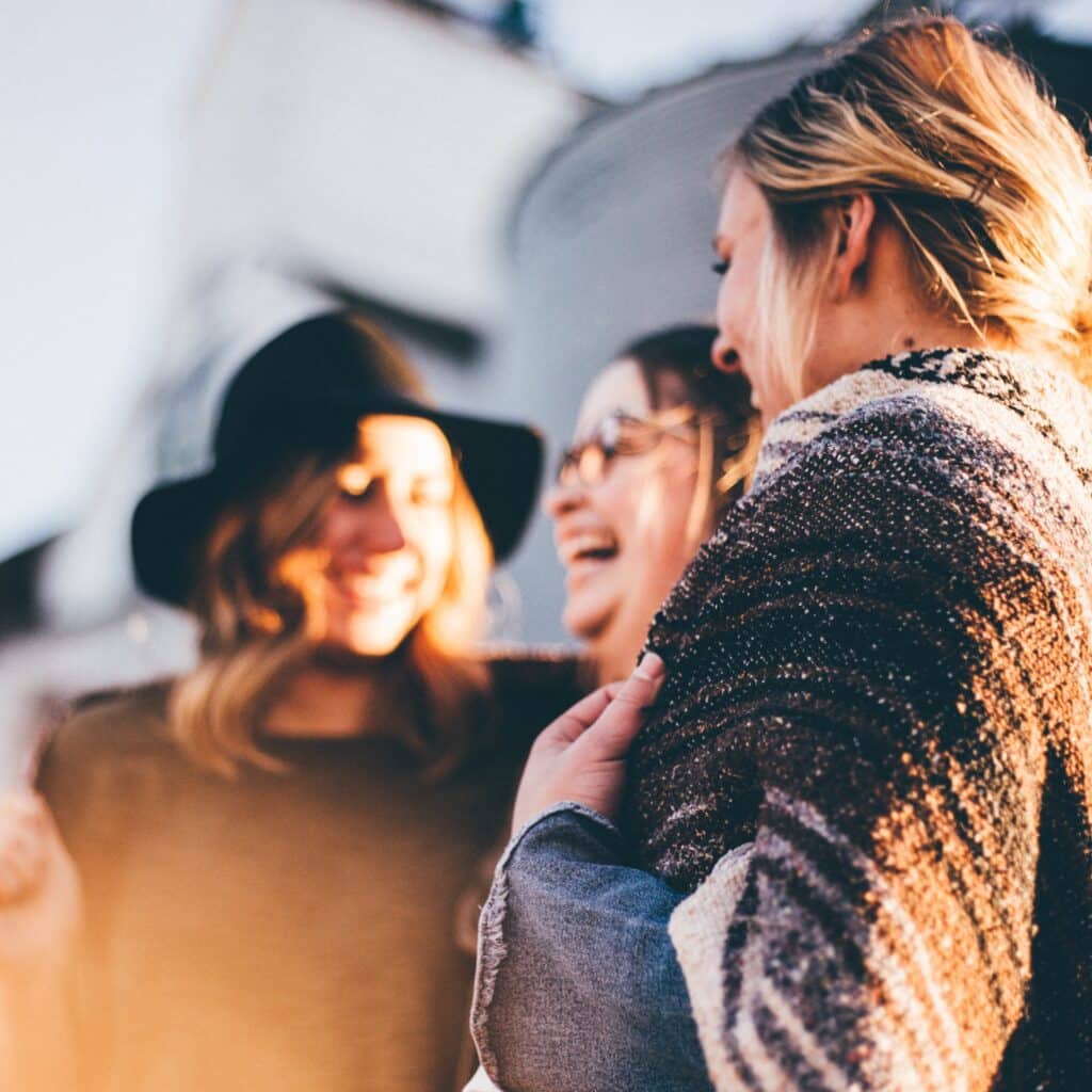 Three women laughing and enjoying each other's company outdoors, with one wearing a hat and the others wrapped in blankets.