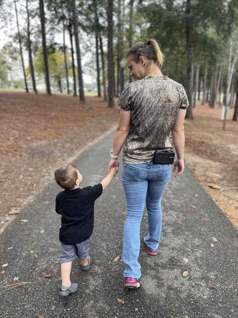 A woman and a child walk hand in hand along a tree-lined path in a park, with the woman wearing jeans and the child in a black shirt.