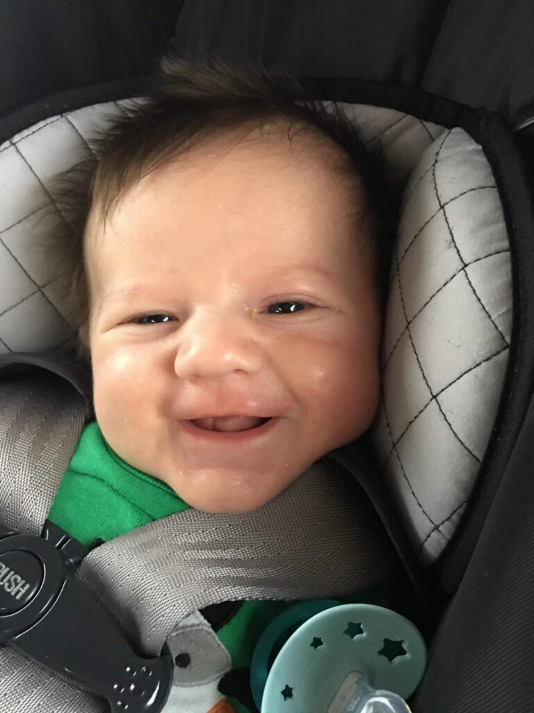 A baby with tousled hair smiles while sitting in a car seat, wearing a green outfit and holding a green pacifier with a star pattern.