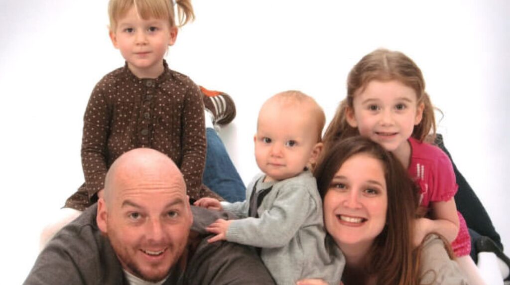 A family of five is lying on a white background, with parents in front and three young children stacked on top. The group is smiling.