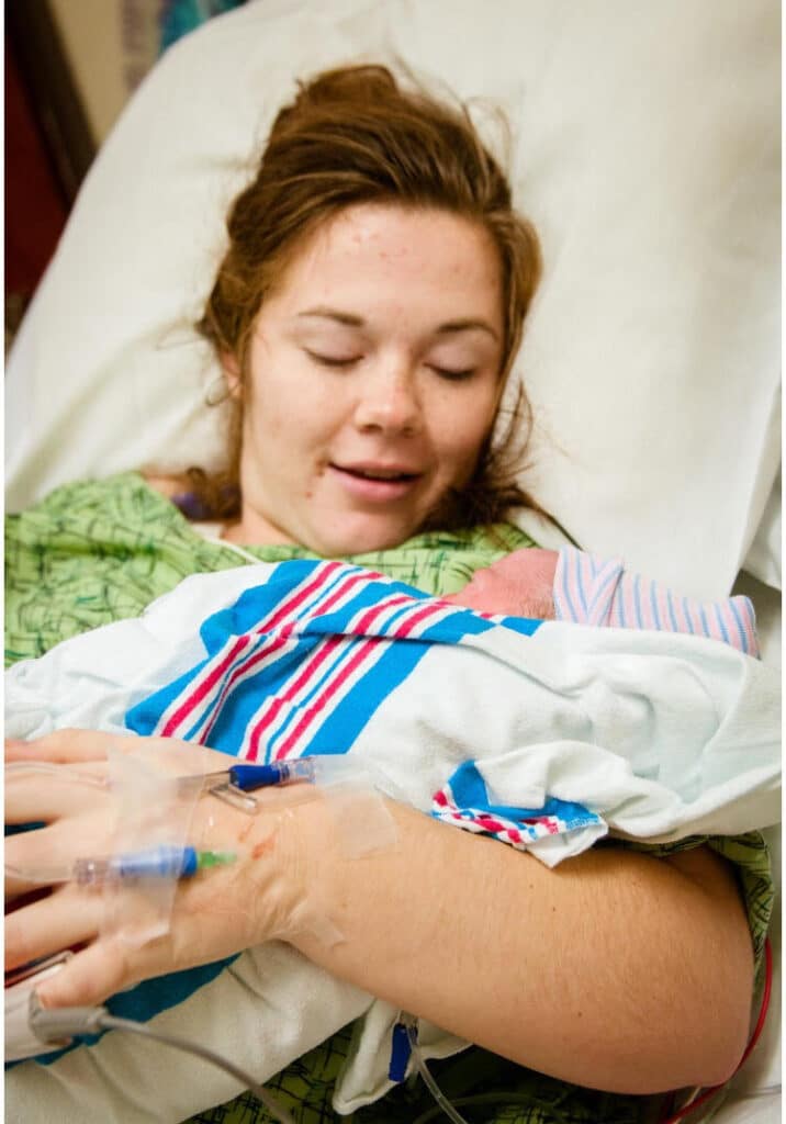 A woman lies on a hospital bed, holding a newborn wrapped in a blanket, with a medical IV in her hand.