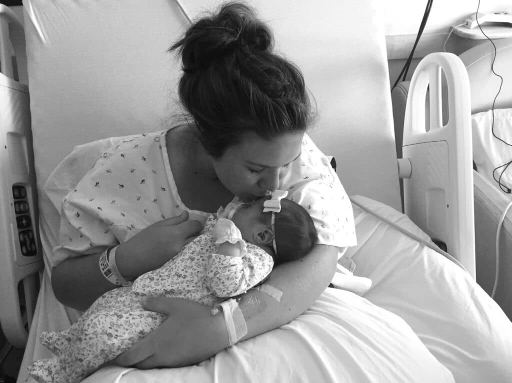 A mother in a hospital bed gently kisses her newborn baby, who is swaddled in a floral outfit. Both are resting peacefully.