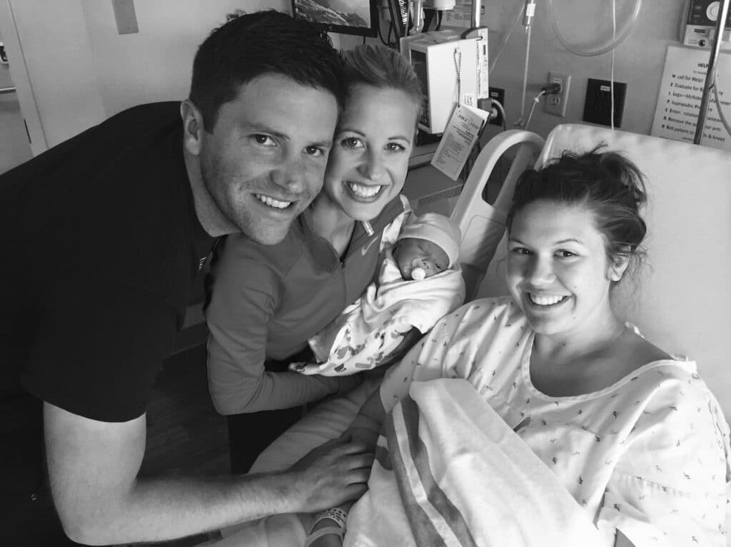 A joyful group celebrates a newborn in a hospital room. A woman holds the baby while another woman and a man smile beside them.