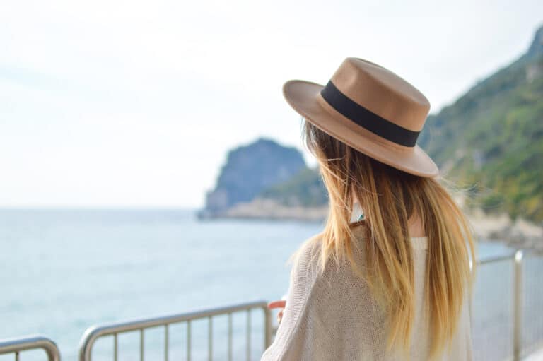 A woman wearing a wide-brimmed hat looks out at the ocean from behind a metal railing, with cliffs visible in the background.