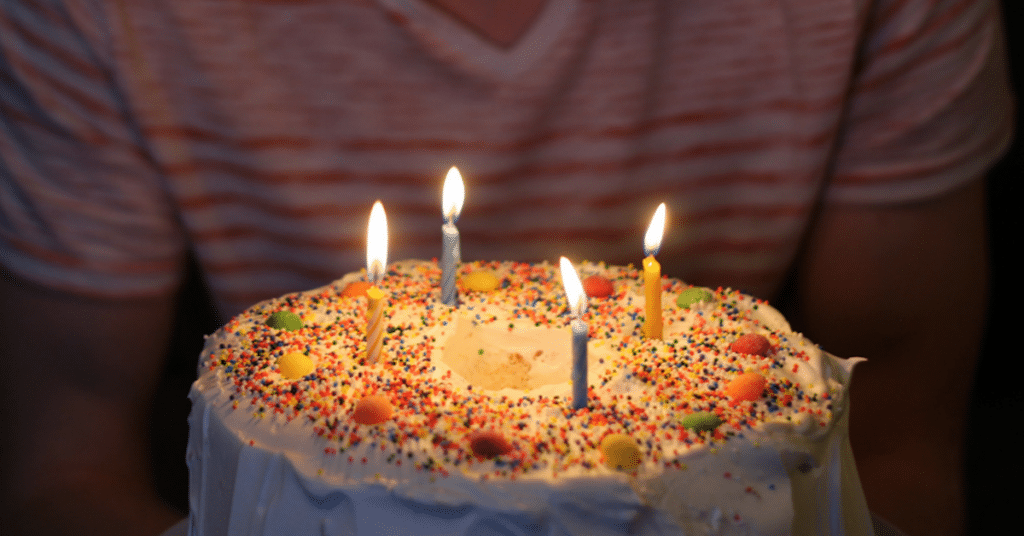 A person holding a birthday cake with five lit candles. The cake is covered in white frosting and colorful sprinkles.