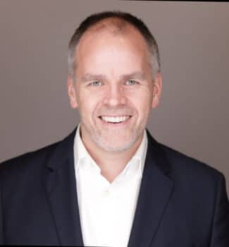 A man with short gray hair and a beard smiles, wearing a dark suit jacket and white shirt. He stands against a plain background.