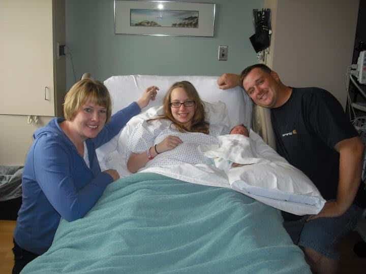 A woman in a hospital bed holding a newborn is surrounded by three smiling adults in a hospital room.