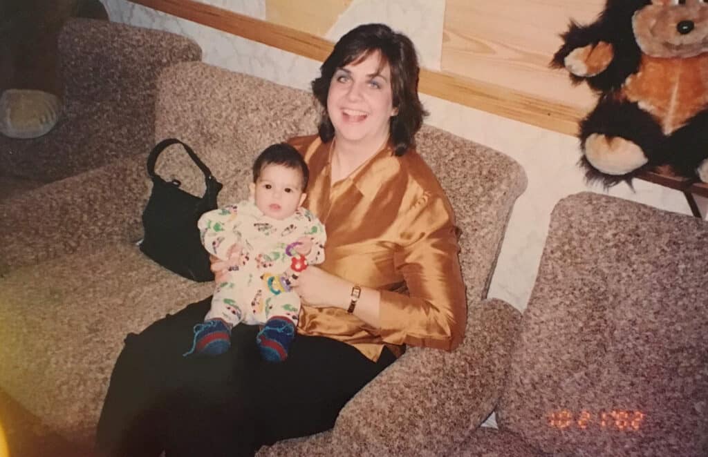 A woman smiles while sitting on a couch holding a baby in her lap. A stuffed animal is visible in the background.