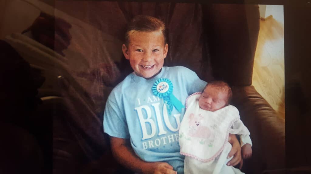 A smiling young boy wearing a "Big Brother" shirt holds a newborn baby wrapped in a blanket on a chair.