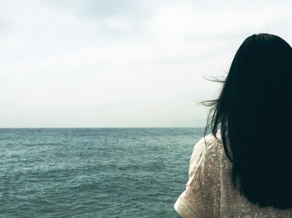 A person with long dark hair stands by the ocean, looking at the water under a cloudy sky.