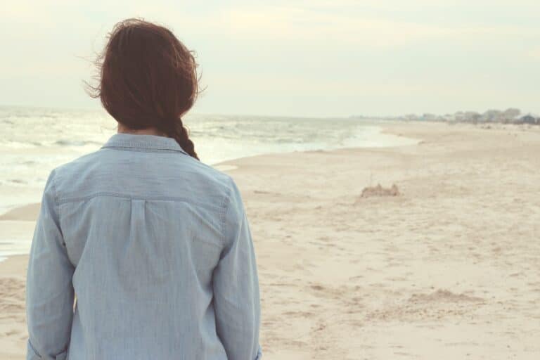 A person in a denim shirt stands on a sandy beach, facing the ocean on a cloudy day.