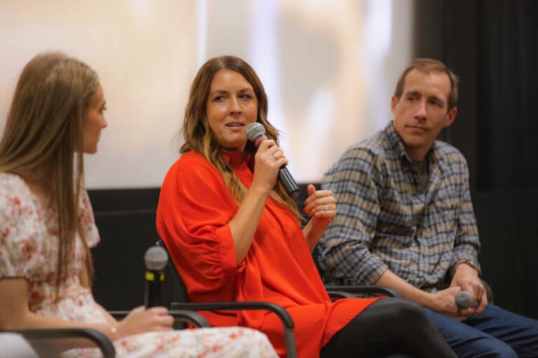 Three people sitting and engaging in a discussion. The woman in the center, dressed in a red top, is speaking into a microphone.