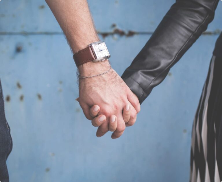 Two people holding hands, one wearing a leather jacket and the other a watch, against a blue, slightly weathered background.