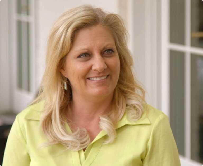 Woman with long blonde hair smiling, wearing a light green shirt, standing in front of a white building with large windows.