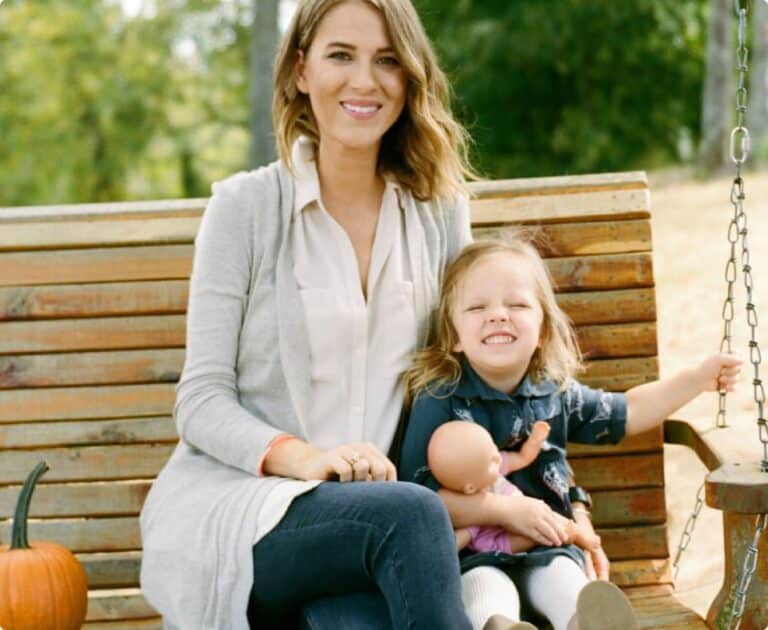 Woman and child sitting on a wooden swing outdoors. The child holds a doll, and there's a small pumpkin next to them. Both are smiling.