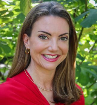 A woman with long brown hair, wearing a red top and gold hoop earrings, smiling in front of a green leafy background.