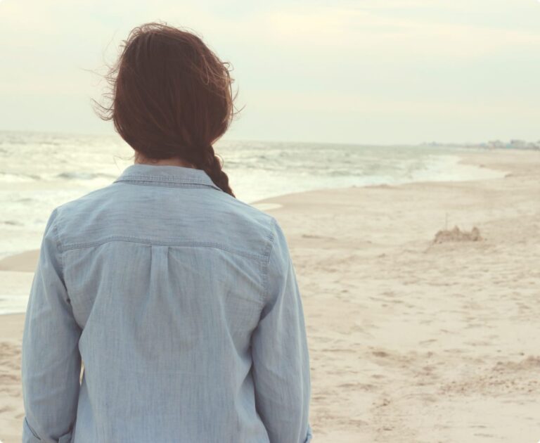 Person with a braid wearing a blue shirt facing the ocean on a calm, empty beach.