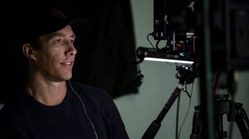 A person wearing a hat and headphones is sitting next to a camera setup, looking intently at something off-screen.