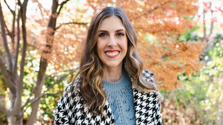 A woman with long wavy hair smiles brightly, wearing a houndstooth coat and blue sweater, surrounded by autumn foliage.