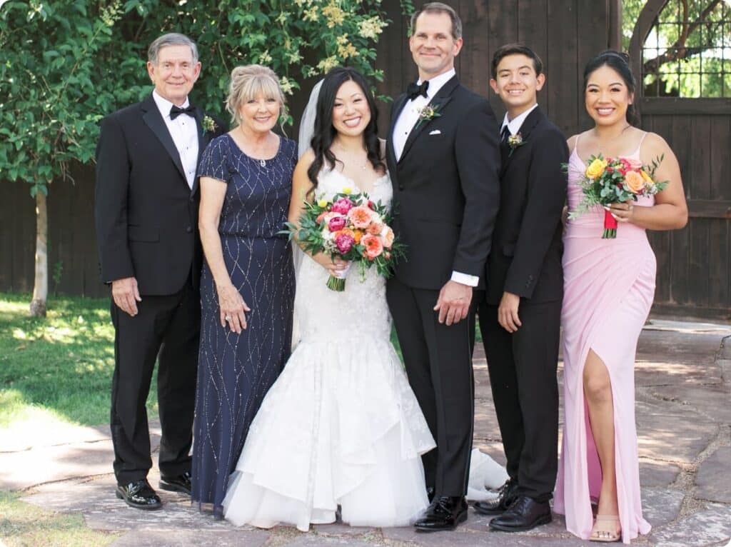 A wedding party, some in black suits and some in formal dresses, stand together smiling and holding bouquets in an outdoor setting.