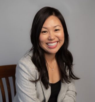 A woman with long dark hair and a big smile, wearing a light gray blazer and black top, sits on a wooden chair against a gray background.