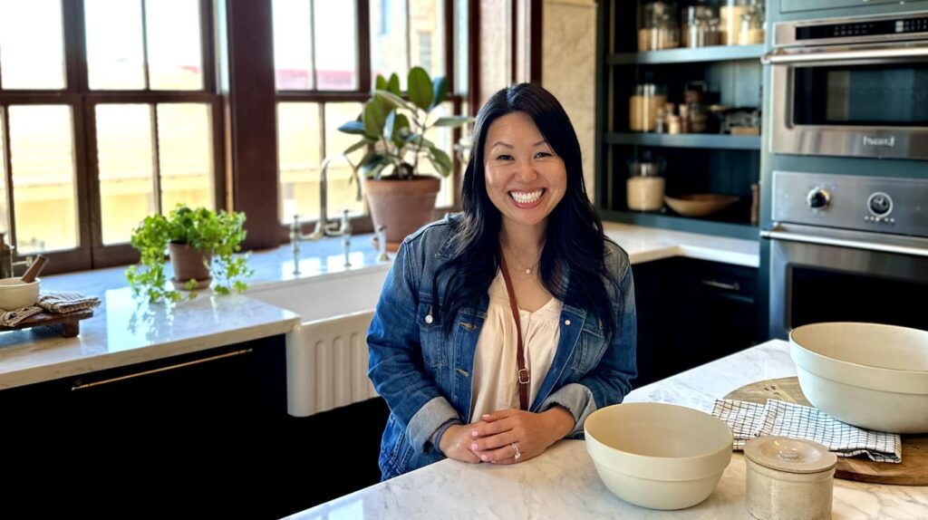 A woman smiles while standing in a modern kitchen with a large window, surrounded by bowls and potted plants.