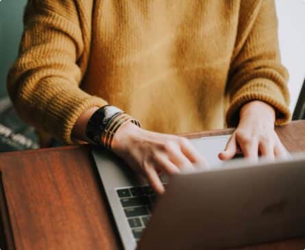 Person in a yellow sweater typing on a laptop at a wooden table, wearing a black bracelet with gold bangles on the wrist.